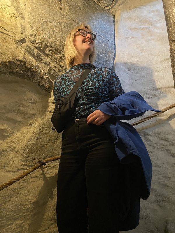 A person standing indoors against a stone wall wearing a small black crossbody bag, holding a jacket over one arm.