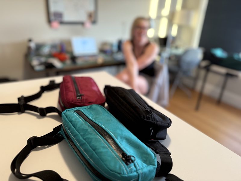Three small zippered bags on a table in the foreground, with a person sitting out of focus in the background of a living space.